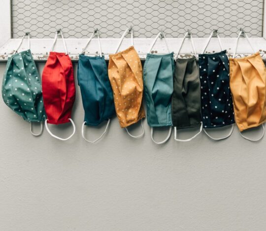 Eight colorful fabric face masks with different patterns hang side by side on a wire mesh frame against a light gray wall.