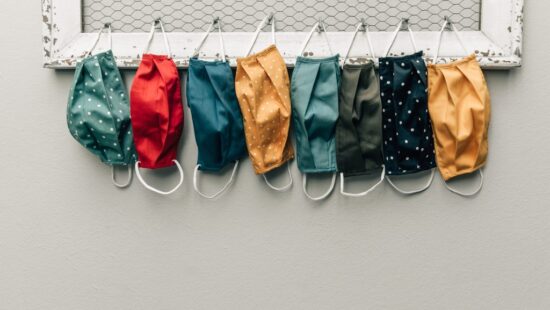 Eight colorful fabric face masks with different patterns hang side by side on a wire mesh frame against a light gray wall.