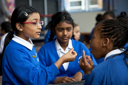 Three students in blue uniforms are engaged in a sign language conversation. One student is signing while the others attentively watch. The background shows a classroom setting.