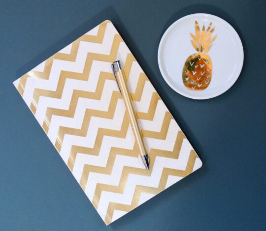 A notebook with a gold chevron pattern and a pen rest on a blue surface next to a small white dish with a gold pineapple design.