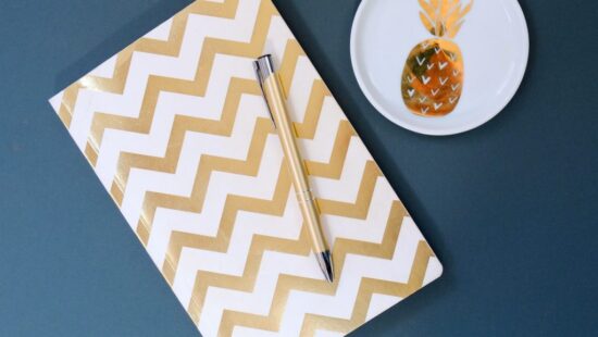 A notebook with a gold chevron pattern and a pen rest on a blue surface next to a small white dish with a gold pineapple design.