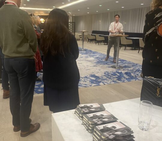 A group of people stands listening to a person speaking in an office setting. Books are stacked on a table in the foreground.