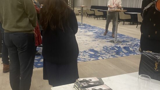 A group of people stands listening to a person speaking in an office setting. Books are stacked on a table in the foreground.