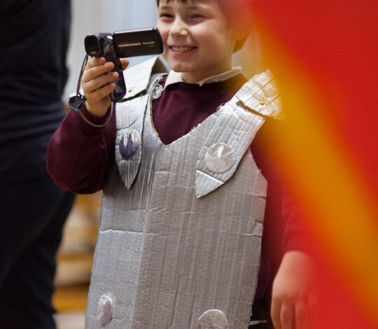 A child wearing a homemade cardboard armor costume holds and looks through a camcorder, smiling.