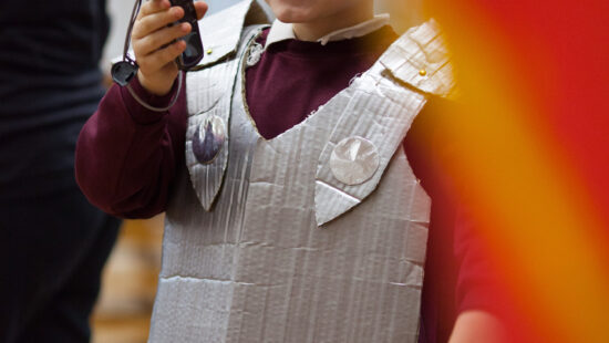 A child wearing a homemade cardboard armor costume holds and looks through a camcorder, smiling.