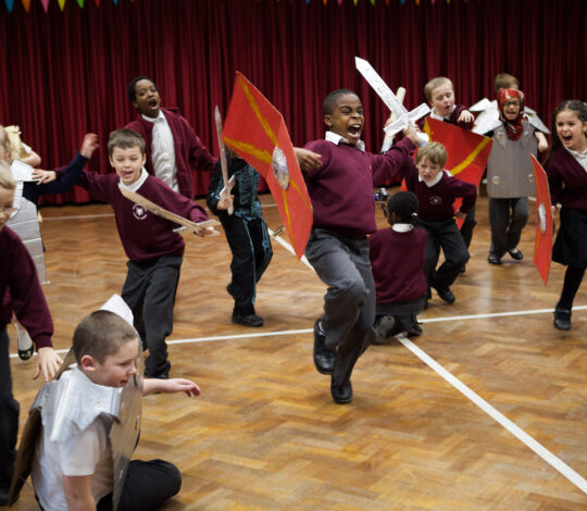 A group of children in matching uniforms engage in a spirited medieval-themed activity, wielding toy swords and shields, in a gymnasium with red curtains and wooden flooring.