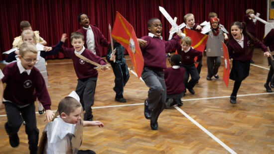 A group of children in matching uniforms engage in a spirited medieval-themed activity, wielding toy swords and shields, in a gymnasium with red curtains and wooden flooring.