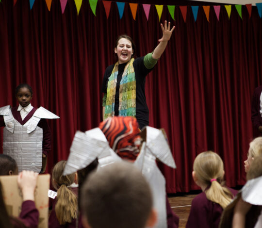 A woman stands on stage in front of a red curtain, gesturing with one hand raised. Children in costume and school uniforms are on stage and in the audience. Multicolored bunting is above.