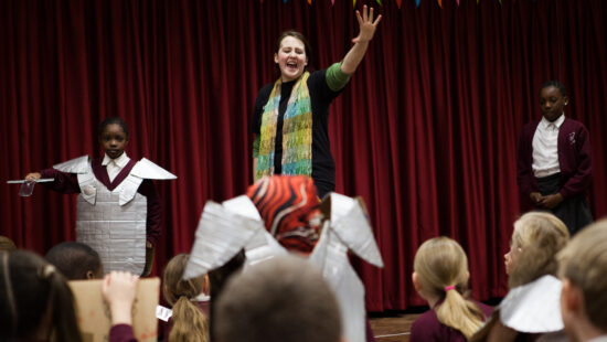 A woman stands on stage in front of a red curtain, gesturing with one hand raised. Children in costume and school uniforms are on stage and in the audience. Multicolored bunting is above.