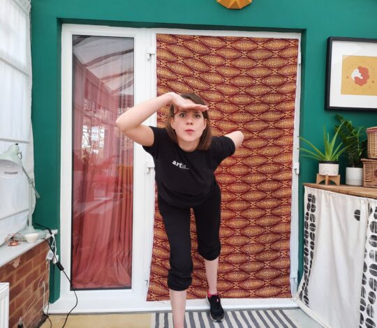 A person in black athletic wear stands on one leg and reaches forward with both arms. They are indoors, in front of a green wall and a door with a decorative curtain. Various items are on a nearby table.