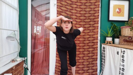 A person in black athletic wear stands on one leg and reaches forward with both arms. They are indoors, in front of a green wall and a door with a decorative curtain. Various items are on a nearby table.