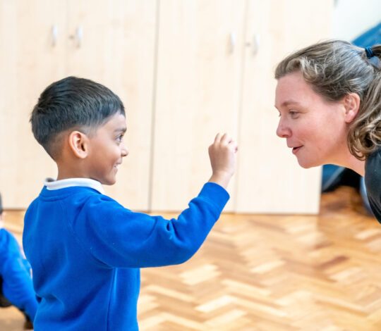 A child wearing a blue sweater smiles and interacts with an adult bending down at his level in a classroom. Another child is partially visible in the background.