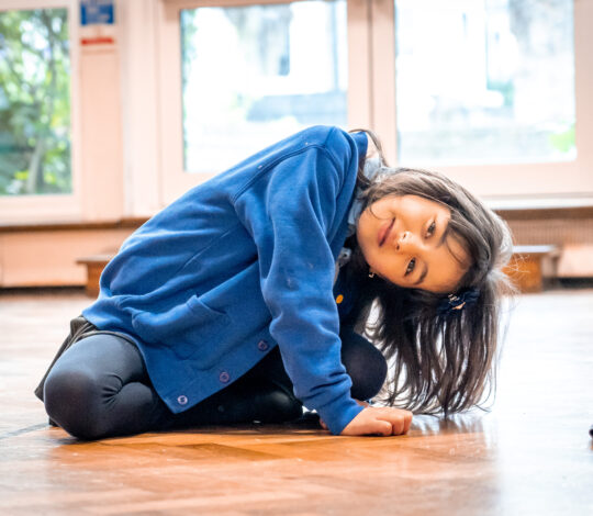 A young girl in a blue jacket sits on the wooden floor, leaning to one side, with a curious expression. Large windows in the background let in natural light.