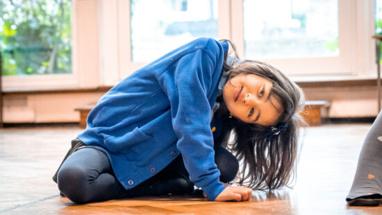 A young girl in a blue jacket sits on the wooden floor, leaning to one side, with a curious expression. Large windows in the background let in natural light.