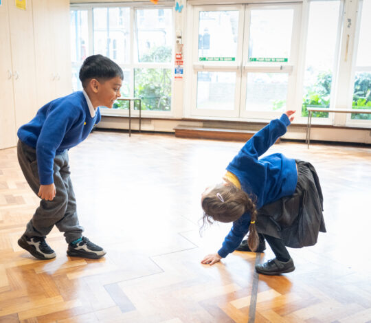 Two young children in school uniforms engage in playful movement on a wooden floor in a brightly lit room.