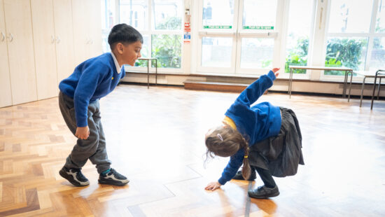 Two young children in school uniforms engage in playful movement on a wooden floor in a brightly lit room.