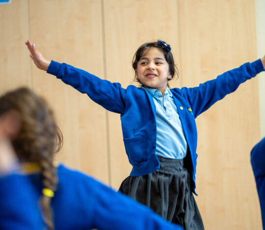 A young girl, wearing a blue cardigan and grey skirt, smiles with her arms stretched out in a classroom setting. She appears to be engaged in an interactive activity with other children.