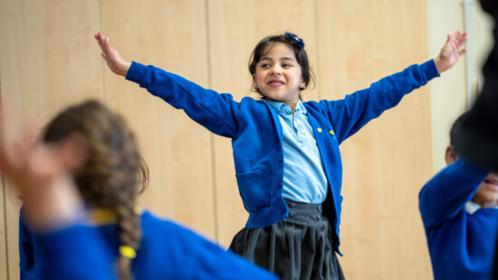 A young girl, wearing a blue cardigan and grey skirt, smiles with her arms stretched out in a classroom setting. She appears to be engaged in an interactive activity with other children.