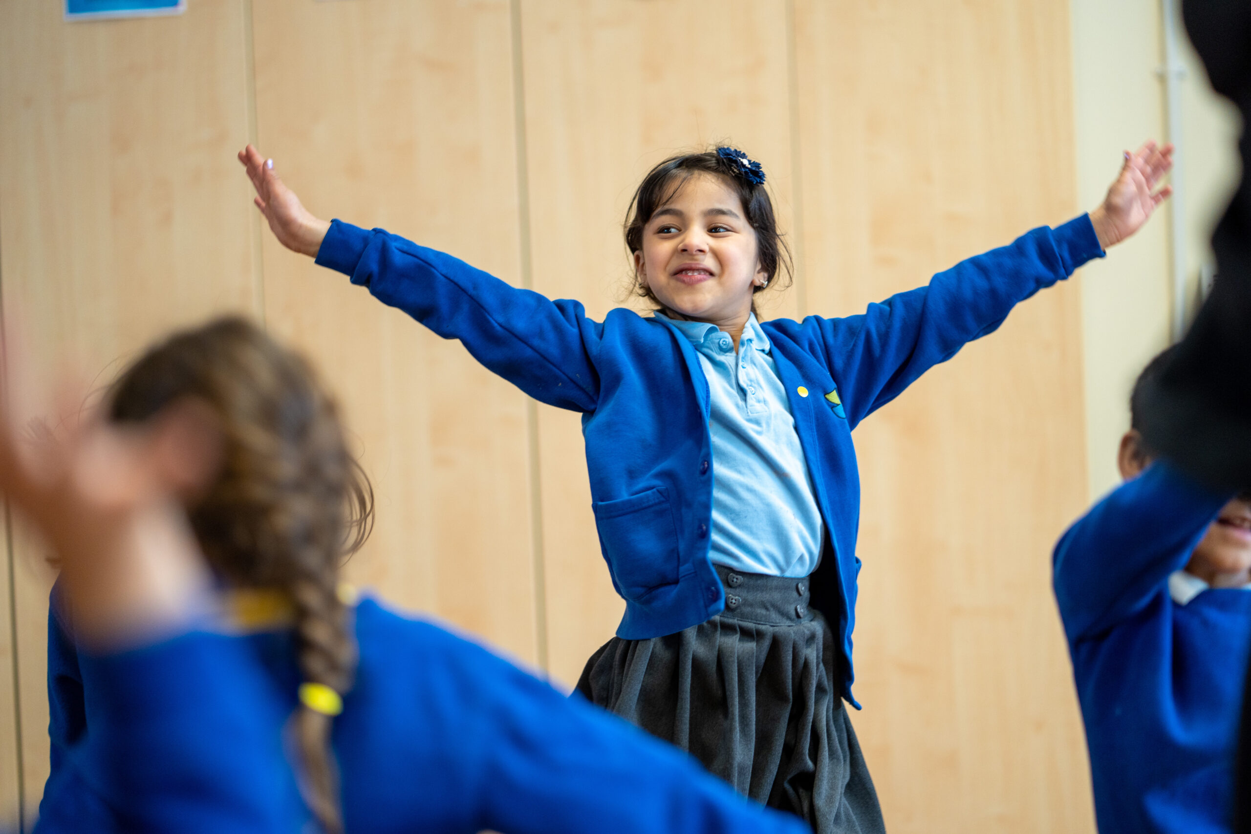 A young girl, wearing a blue cardigan and grey skirt, smiles with her arms stretched out in a classroom setting. She appears to be engaged in an interactive activity with other children.