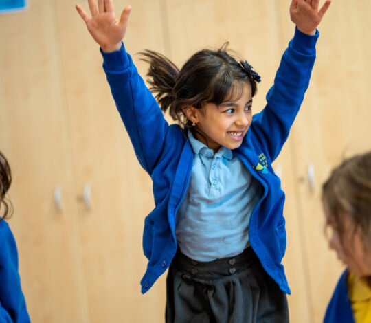 A young girl in a blue school uniform raises her arms enthusiastically, standing indoors with other children.