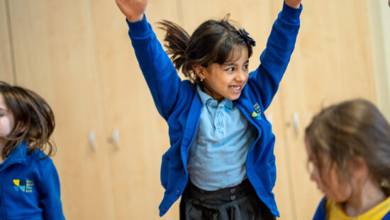 A young girl in a blue school uniform raises her arms enthusiastically, standing indoors with other children.