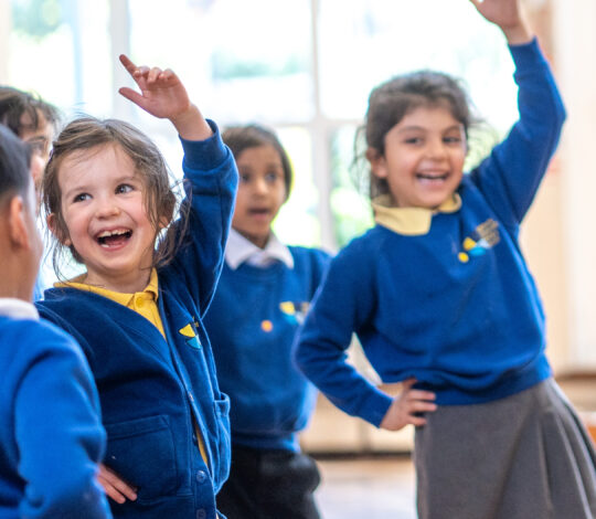 A group of children in blue school uniforms are enthusiastically participating in a classroom activity, with some raising their hands and smiling.