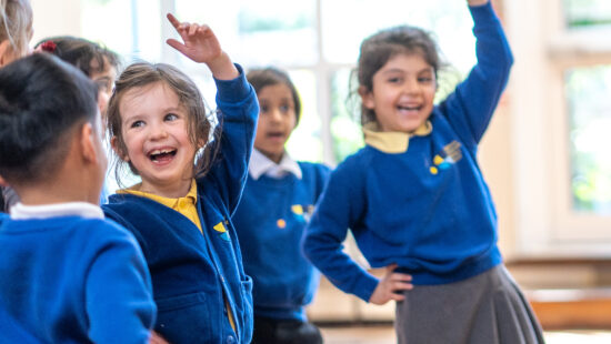 A group of children in blue school uniforms are enthusiastically participating in a classroom activity, with some raising their hands and smiling.
