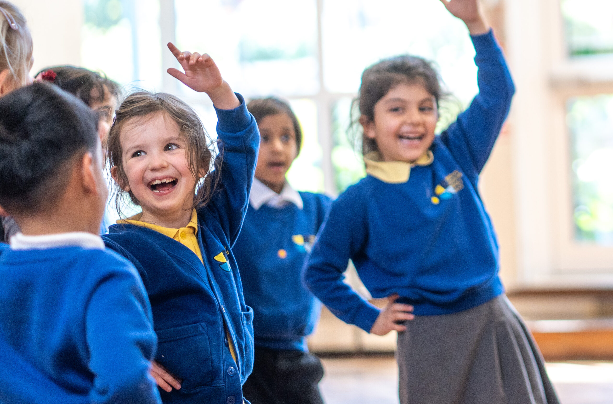 A group of children in blue school uniforms are enthusiastically participating in a classroom activity, with some raising their hands and smiling.