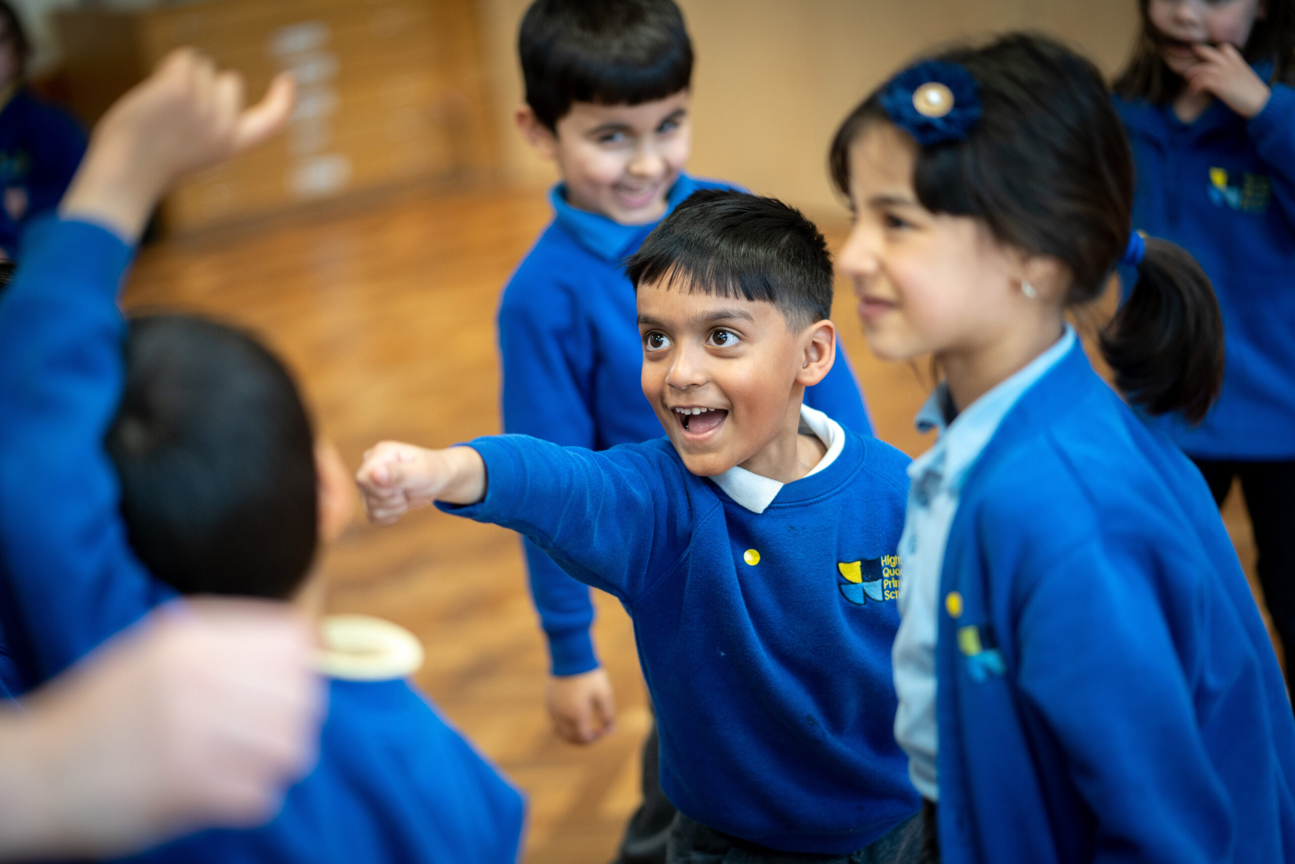 Children in blue school uniforms are engaging in an activity indoors, with one child in the center pointing excitedly and others nearby smiling and interacting.