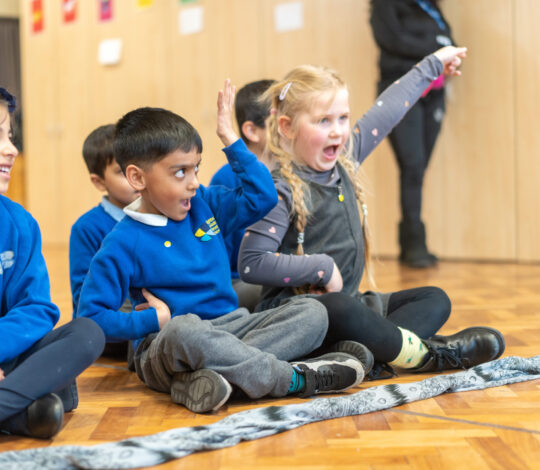 Children sitting on the floor in a classroom, with one child raising their hand and another expressing excitement.