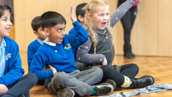 Children sitting on the floor in a classroom, with one child raising their hand and another expressing excitement.