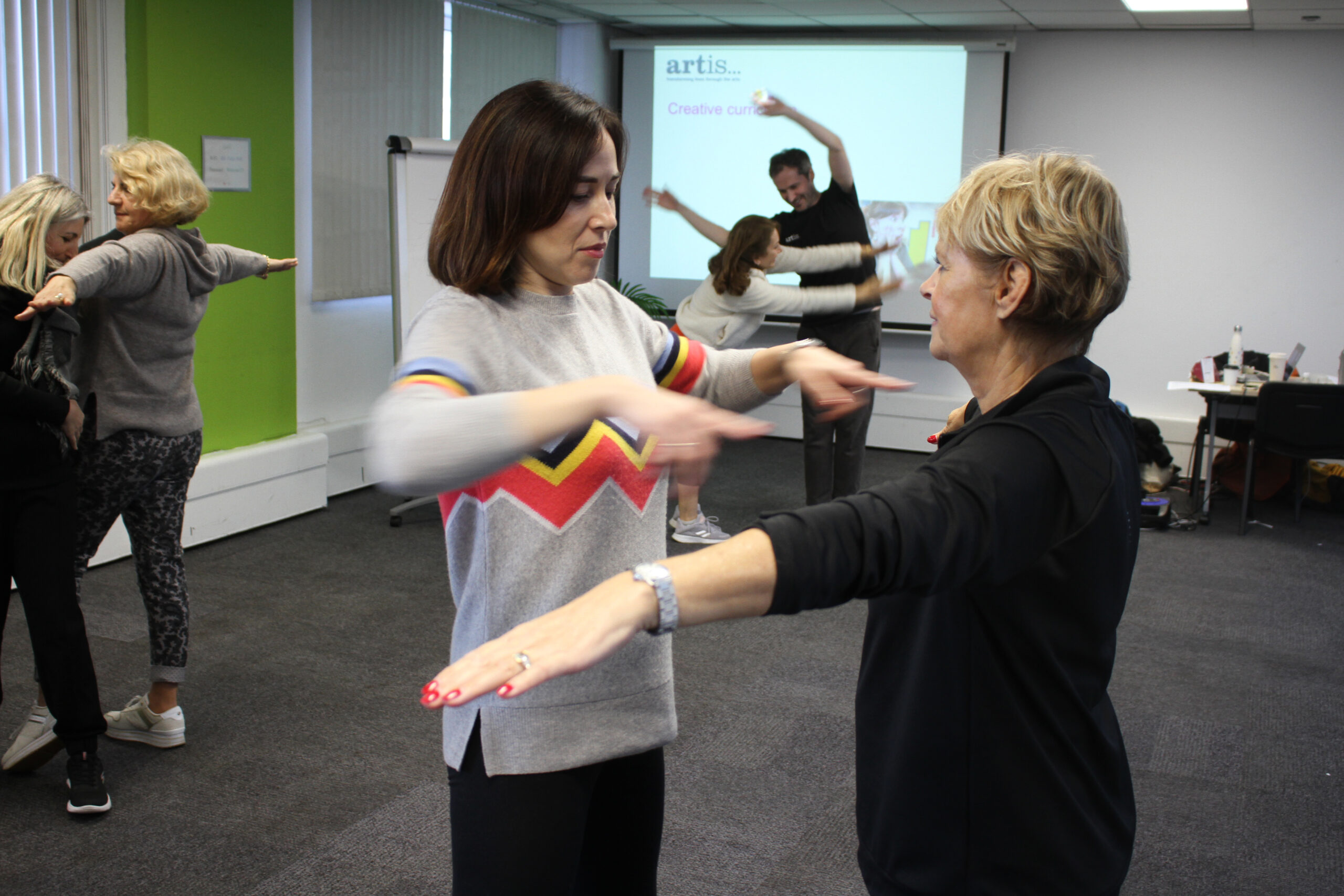 Two pairs of people engage in a guided physical activity inside a classroom, with arms extended and facing each other. A presentation screen and whiteboard are visible in the background.