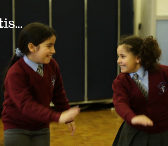 Children in school uniforms engage in a dance or movement activity. Text on the left side reads "artis...".