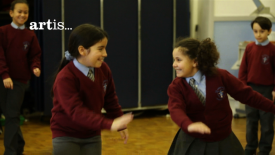 Children in school uniforms engage in a dance or movement activity. Text on the left side reads "artis...".