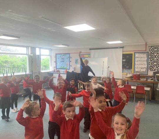 A group of young children wearing red sweaters joyfully raise their hands in a classroom while an adult stands at the front, smiling.