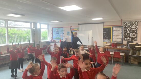 A group of young children wearing red sweaters joyfully raise their hands in a classroom while an adult stands at the front, smiling.