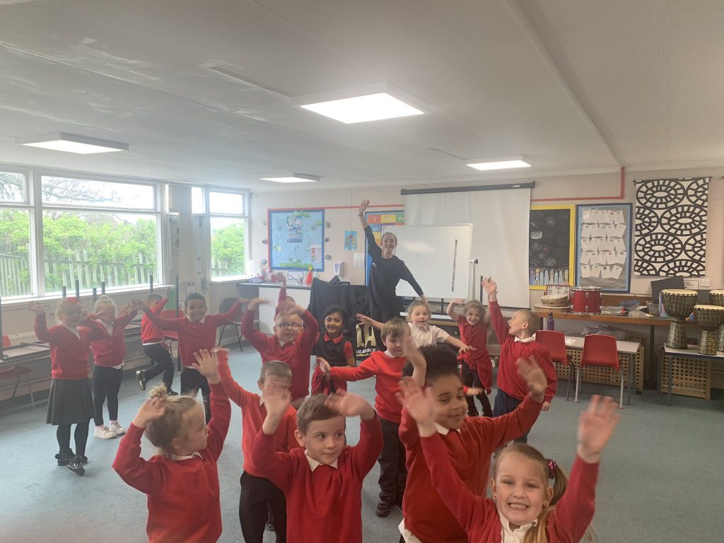 A group of young children wearing red sweaters joyfully raise their hands in a classroom while an adult stands at the front, smiling.