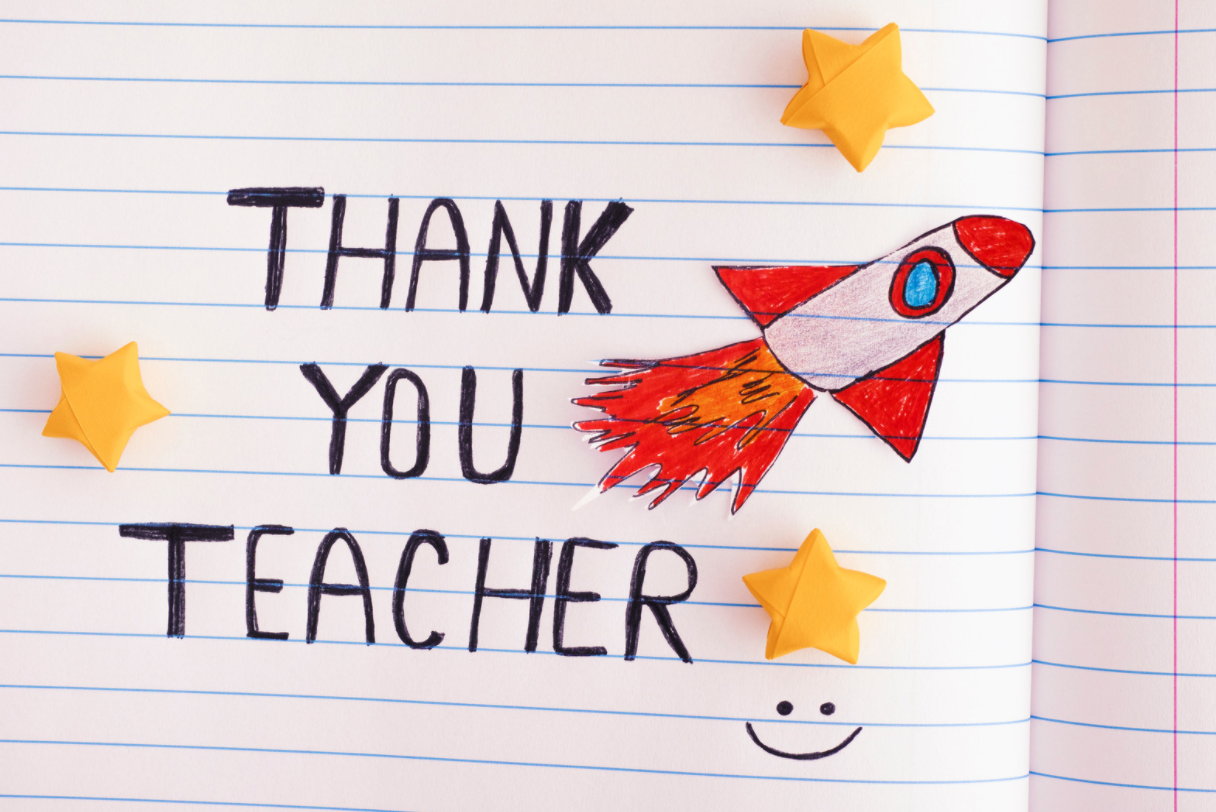 An open notebook with "Thank You Teacher" written in black letters, a red and white rocket drawing, three yellow paper stars, and a small smiley face drawn at the bottom.