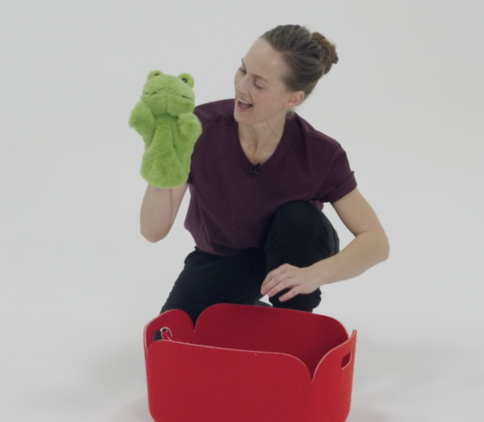 A woman in a purple shirt kneels beside a red storage bin, holding and looking at a green frog puppet, against a plain white background.