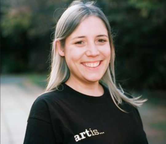 A woman with long hair smiling, wearing a black shirt with the word "artis..." printed on it, standing outdoors. In the background, you can almost hear the onomatopoeic sounds of birds chirping and leaves rustling in the gentle breeze.