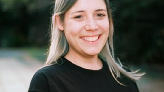 A woman with long hair smiling, wearing a black shirt with the word "artis..." printed on it, standing outdoors. In the background, you can almost hear the onomatopoeic sounds of birds chirping and leaves rustling in the gentle breeze.