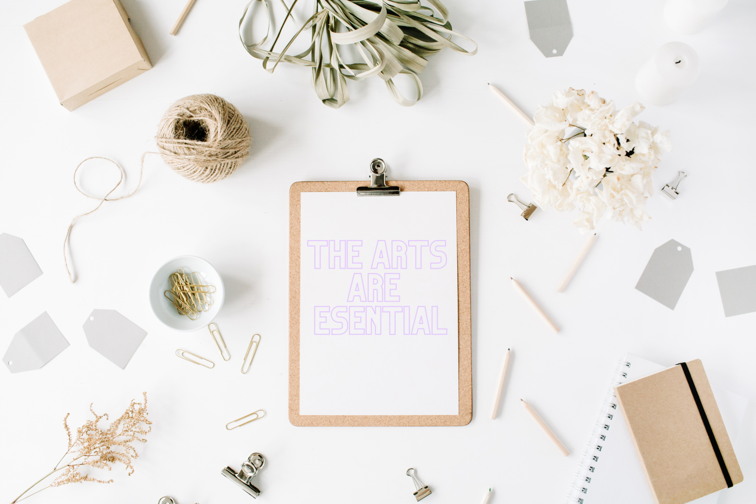 A clipboard with a paper reading "The Arts Are Esential" surrounded by various office and craft supplies on a white surface.