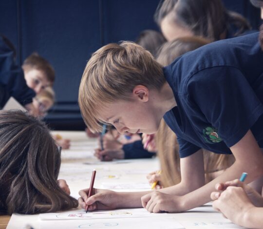 Children in a classroom, wearing dark blue shirts, are intently drawing on paper with colored pencils. They are gathered closely around a large table, concentrating on their artwork.