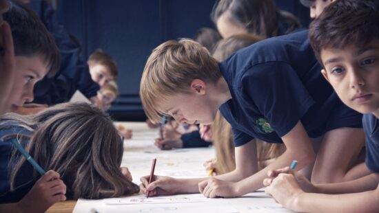 Children in a classroom, wearing dark blue shirts, are intently drawing on paper with colored pencils. They are gathered closely around a large table, concentrating on their artwork.