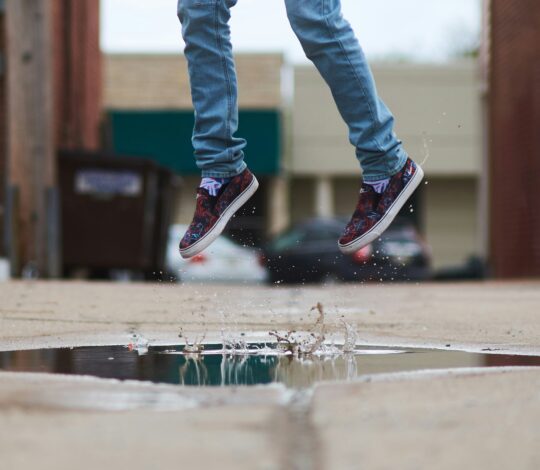 A person in jeans and colorful sneakers is jumping over a puddle in an alley, with water splashing below.