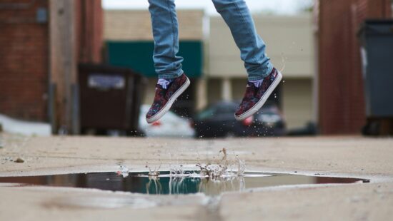 A person in jeans and colorful sneakers is jumping over a puddle in an alley, with water splashing below.