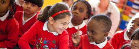 Children wearing red school uniforms sit together in a classroom, with two of them smiling and engaged in conversation.