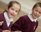 Two young children wearing maroon school uniforms and white shirts, smiling and looking slightly to the side. The background is slightly blurred.