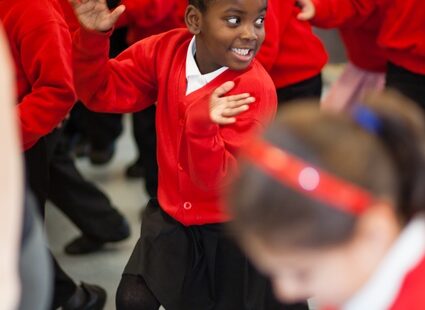 A group of young children in red sweaters participate in an energetic activity inside a classroom. One girl in the foreground is smiling broadly and appears to be enjoying the activity.