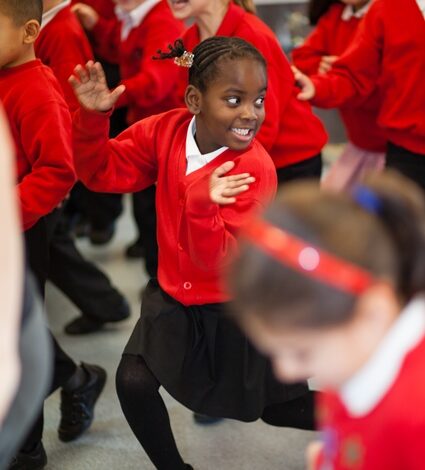 A group of young children in red sweaters participate in an energetic activity inside a classroom. One girl in the foreground is smiling broadly and appears to be enjoying the activity.